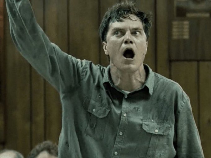 A man with dark hair wearing a dirty grey button-up shirt raises his arm with a shocked, open-mouthed expression against a wood-paneled background.