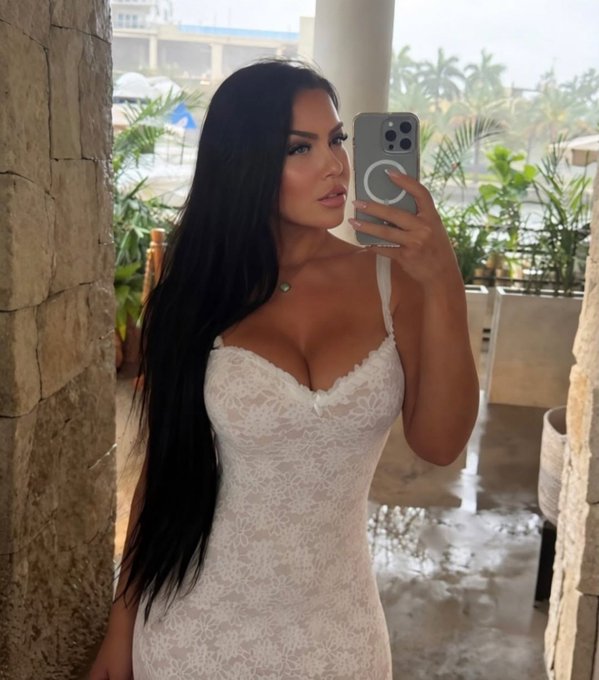A woman with long dark hair taking a mirror selfie with her iPhone while wearing a white lace dress in what appears to be a resort or hotel setting.
