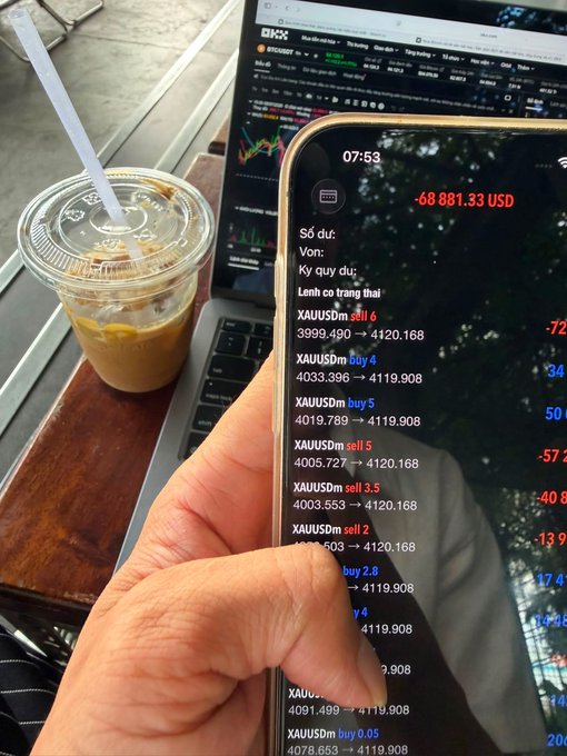 A hand holding a phone showing a trading account with a balance of -68,881.33 USD displaying XAUUSD trading positions (sell 6, buy 4, buy 5, etc.), with an iced coffee and a laptop showing BTC/USDT charts in the background.