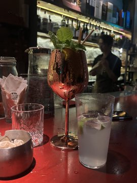 The image shows a bar scene with various drinks and glasses placed on a red table, including a copper-colored wine glass filled with a drink that appears to be a cocktail or mixed drink. There are also several other glasses of different shapes and sizes scattered across the table. A person can be seen in the background, possibly working behind the counter or attending to customers. The image is related to the post "Crypto Survival Plan" as it features a bar scene with drinks and glasses, which could be used for socializing or enjoying leisure time during a break from cryptocurrency trading or investing activities.