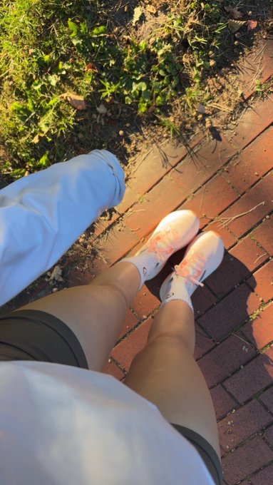 A first-person view photo of someone wearing white sneakers with peach-colored laces and light pants, on a brick path with grass and weeds nearby in sunlight.