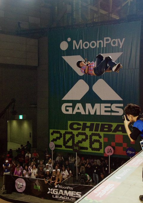 A skateboarder performs an aerial trick in front of a large "MoonPay X Games Chiba 2026" banner at the X Games League event, while a photographer captures the action from the ramp.