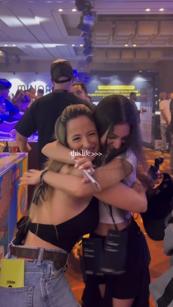 Two young women embrace at an event with the text 'this life >>>' overlaid on the image.