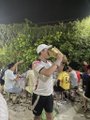 A very small, blurry image showing a group of people outdoors in casual summer clothing, with one person in the center wearing a white t-shirt and a hat posing with a hand gesture.