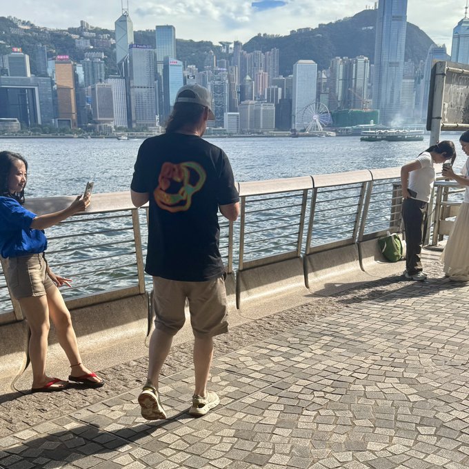 Hello Hong Kong Bored Ape Yacht Club is the username of the person taking this photo from the perspective of a back view of someone. The photo taken appears to be of the Hong Kong skyline from a dock area of the city. There is a inclination to think that this photo was taken inside of a world tour given the number of different people and their poses in this single photo. From the add-on, it seems this visit is a form of tourism or an interest of culture of this location.