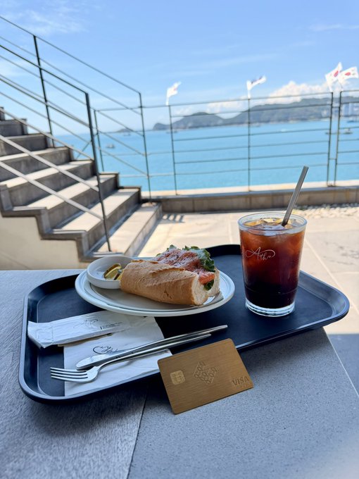 A cappuccino and sandwich meal on a tray overlooks a stunning seaside view, with an "Air" branded cold brew coffee and a credit card visible on the table.

The image showcases a luxurious lifestyle moment—a seaside café meal paired with what appears to be a premium metal credit card—visually reinforcing the post's promotion of EtherFi's upgraded VIP membership benefits, which include perks like free shipping for the Gold Metal card.