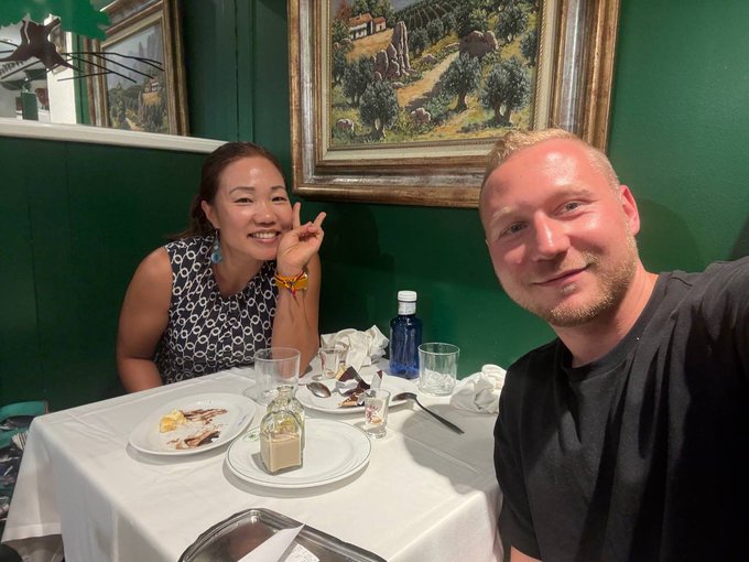 A smiling Asian woman and a man taking a selfie together at a restaurant table set with plates of food, a water bottle, and crumpled napkins against a green wall with framed artwork.