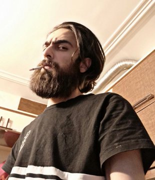 The image depicts a bearded man with a long beard wearing a black shirt. He has a cigarette in his mouth while looking directly at the camera, giving off an intense expression. The image also features a caption that reads "Good morning, lovely community If you're interested in photography, I have several pieces available on @objktcom across different collections, moods, and techniques. Feel free to explore them through the link in my bio". This could be interpreted as an advertisement or recommendation for photography-related content.