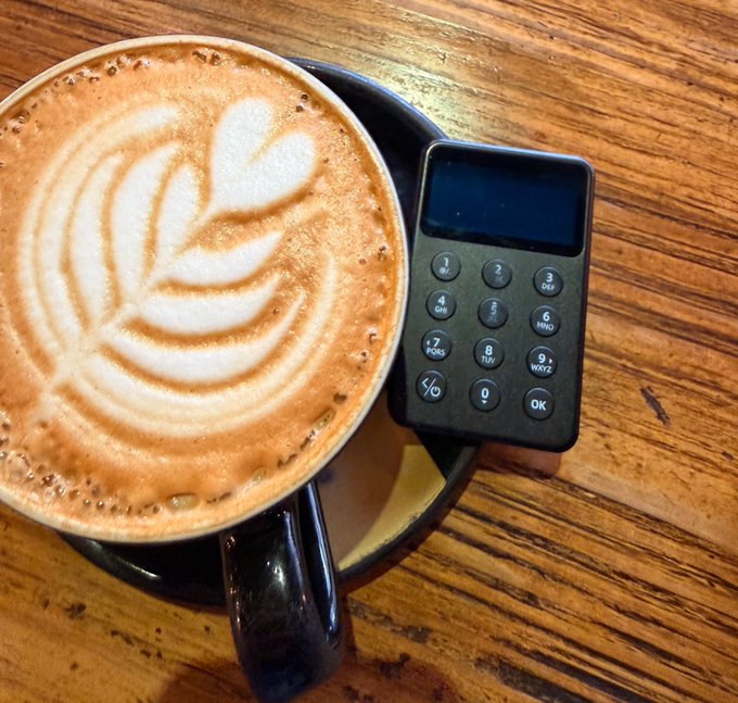 A cappuccino with intricate latte art sits beside a small hardware wallet device featuring a numeric keypad (digits 0-9 plus buttons like "QR", "OK", and letter groups ABC/DEF/GHI/JKL/MNO/PQRS/TUV/WXYZ) on a wooden table—visually pairing the post's "coffee for your health" and "hardware wallet for your wealth" message.