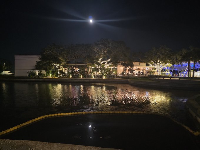 A nighttime scene of a waterfront promenade with illuminated trees and buildings reflected in the calm water under a bright full moon.