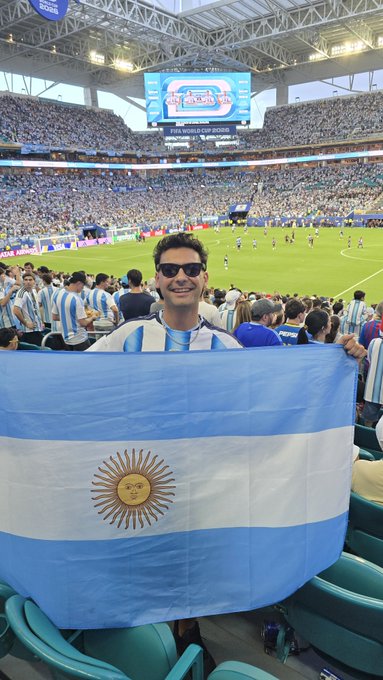 A young man wearing sunglasses holds up an Argentina flag while attending the FIFA World Cup match, visible on the stadium screen in the background.
