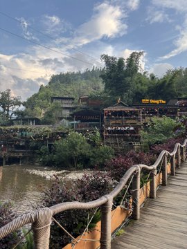 A wooden boardwalk winds along a riverside toward a traditional Chinese-style resort labeled "Kong Shan," set against lush green hills. The caption "imagine getting liquidated here" pairs ironically with the serene, picturesque setting—juxtaposing the peaceful beauty of the location with the harsh reality of financial ruin, suggesting the tranquil scene offers cold comfort to someone facing devastating trading losses.