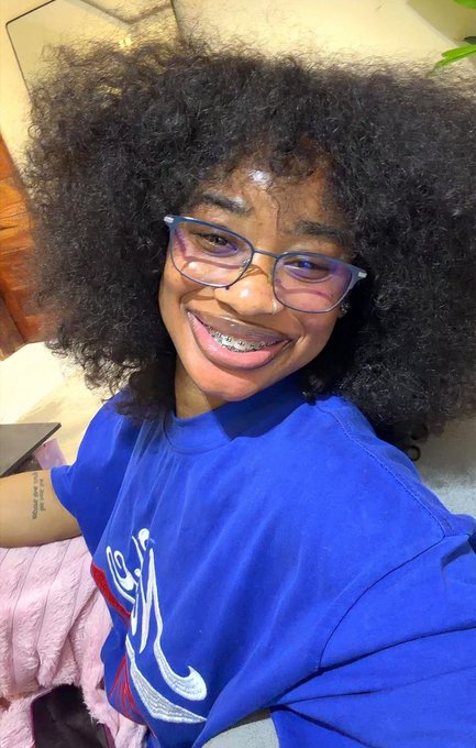 A selfie of a smiling young woman with voluminous curly hair, glasses, and braces, wearing a royal blue t-shirt with a graphic print.