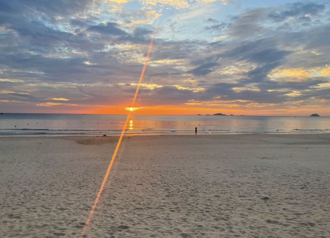 A serene sunset over a beach with a person walking in the shallow water, with a dramatic streak of orange from the setting sun crossing the cloudy sky.