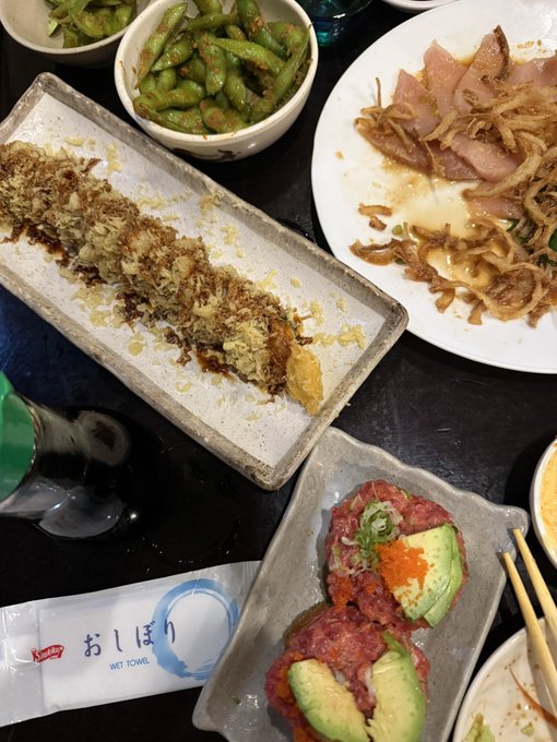A spread of Japanese dishes including edamame, fried items with bonito flakes, raw meat slices with onions, and tuna tartare with avocado, accompanied by a wet towel packet labeled 'おしぼり' (oshibori).