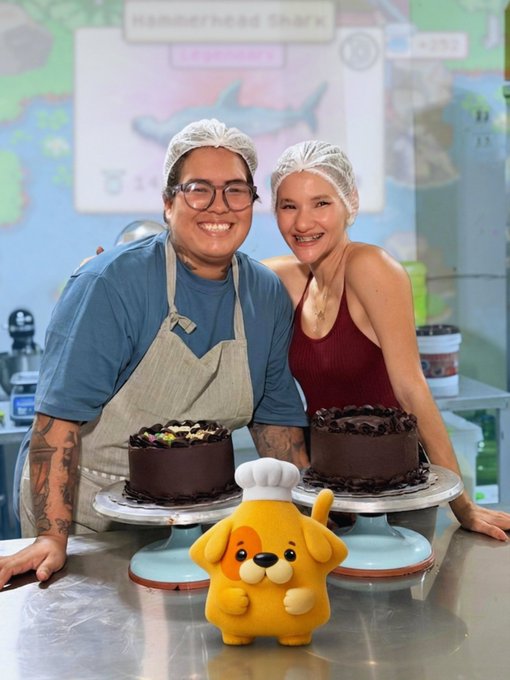 The image features two people posing for a photo, both wearing aprons while standing next to a table filled with cakes. The person on the left has a dog figurine sitting beside them, adding a playful touch to the scene. In the background, there is a sign that reads "Happy Mondaaaaaaaaaaay beautiful @Ronin_Network people! Today was an extremely busy day for me IRL, but despite everything, I got invited to a bakery workshop in my city and ended up making a really cute cake hahaha And yes... @KanstarWorld came with me. Can you believe?". The focus of the image is on the two individuals and their interaction with the cakes, creating an engaging and visually appealing scene that complements the post's content.
