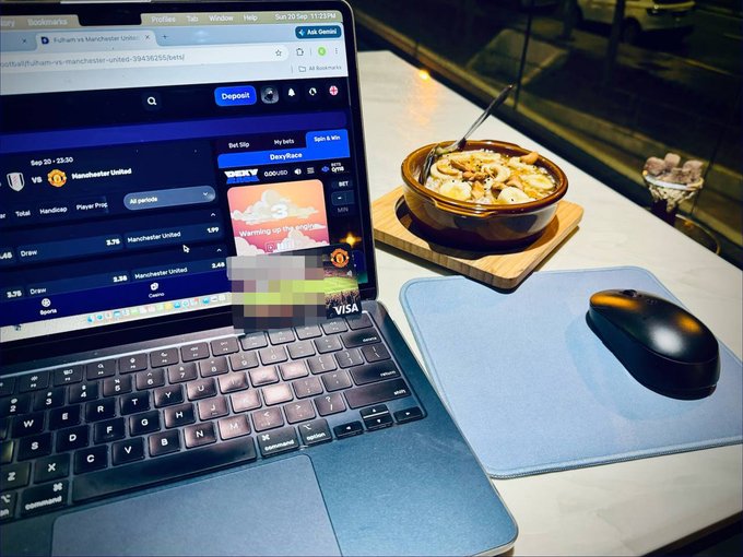 A laptop displaying a sports betting website showing a Manchester United vs. Bournemouth match sits next to a bowl of food and a wireless mouse on a desk.