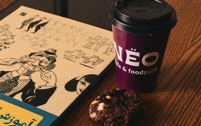 A purple NEO Cafe & Foodpack coffee cup sits on a wooden table next to a chocolate cookie and a notebook with illustrated characters, with Arabic text visible on a book at the bottom left.