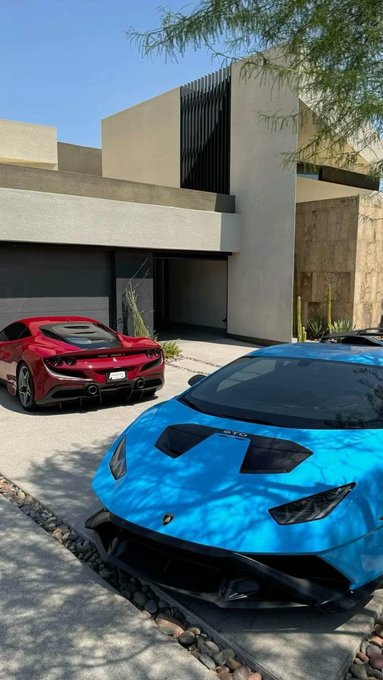 Two luxury sports cars, a red Ferrari and a bright blue Lamborghini, parked in the driveway of a modern minimalist desert home with cacti landscaping.