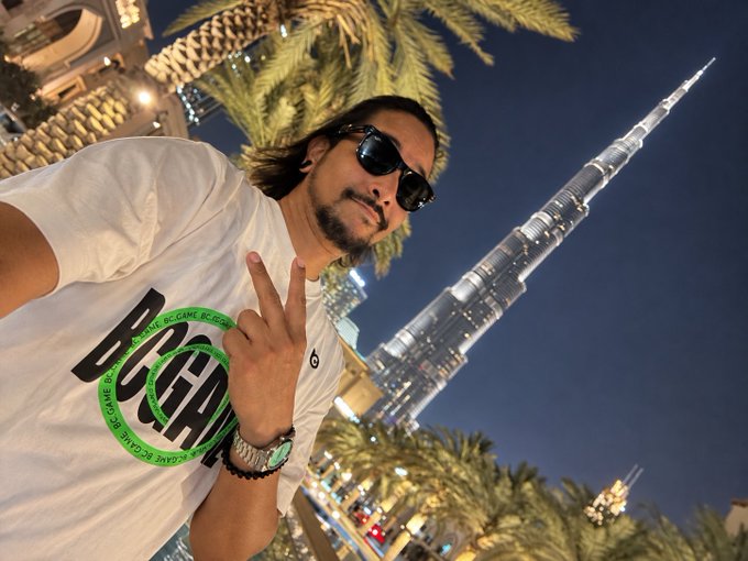 A man wearing sunglasses and a white t-shirt flashes a peace sign in front of the Burj Khalifa at night, with a green "GAME BC.GAME" watermark across his chest. The "Gn degens" caption pairs with the late-night Dubai selfie to bid goodnight to fellow crypto enthusiasts.