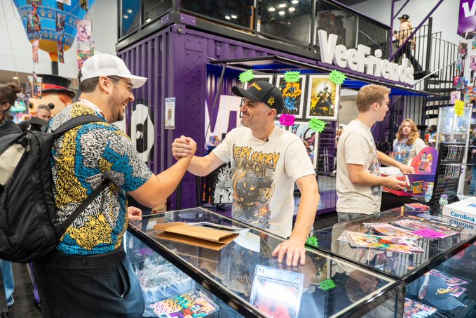 Two men shake hands across a glass display counter at a Veefriends booth at what appears to be a convention or trading card expo.