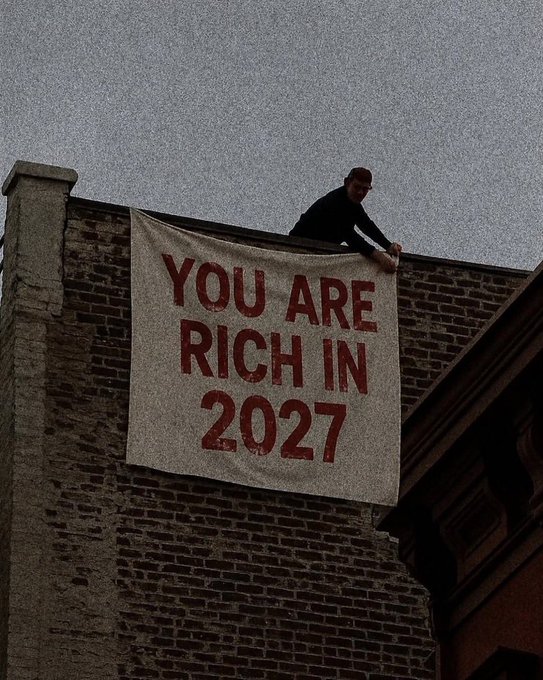 A person on a rooftop unfurls a large banner reading "YOU ARE RICH IN 2027," echoing the post's "Claim it" message by presenting a bold, prophetic affirmation of future prosperity.