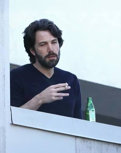 A bearded man with dark hair and a black t-shirt leans on a balcony railing, smoking a cigarette with a green bottle beside him, his expression weary and contemplative. The caption "When you've been locked in the entire year but haven't made millions yet" humorously captures the look of someone whose grind year has yet to pay off.