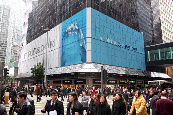 The image shows a busy city street with many people walking around, some of whom are carrying backpacks or handbags. The scene takes place near a tall building that has an advertisement for a cryptocurrency wallet displayed on its side. This advertisement highlights the convenience and accessibility of Bitcoin as a payment method, encouraging users to consider using it instead of traditional banking systems. The image also includes text promoting the use of Bitcoin wallets, which can be seen in various parts of the frame.