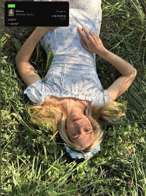 A young woman with blonde hair in pigtails lies upside down in a field of green grass and clover, wearing a white floral sundress. The Spotify overlay shows "Silence" by The One, The O... was added 1 hour ago, matching the caption's theme of gratitude to @jayray7_eth for purchasing the art.