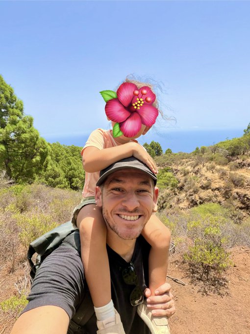 A cheerful selfie of a man with a child on his shoulders during a sunny outdoor hike; the child's face is covered by a digital pink flower sticker.