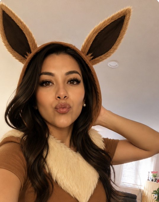 The image features a woman wearing a brown hooded animal head costume, which includes large ears and a tail. She has long hair that falls over her shoulders, giving her a unique appearance. The caption of the photo reads "gQuack Everyone is turning into a Pokémon. @wallchain". This suggests that the woman might be participating in a themed event or party related to Pokémon, possibly as part of a costume contest or a fun gathering with friends and family.

The image also includes a potted plant located near the bottom right corner, adding some greenery to the scene. The overall atmosphere seems lighthearted and playful, with the woman's costume drawing attention and making her stand out in the picture.