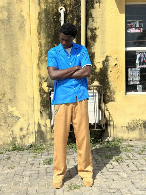 A young man wearing a bright blue short-sleeved shirt, tan pants, and tan boots stands with his arms crossed against a yellow weathered wall.