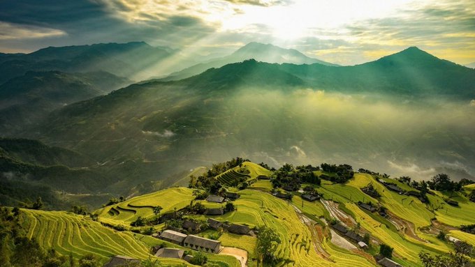 The image shows a beautiful mountainous landscape with a large open field covered in green grass at its center. In the background, there are several houses scattered across the area, adding to the picturesque scenery. The sun shines brightly over the scene, casting light on the mountains and creating a warm glow that illuminates the entire landscape.