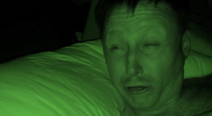 A close-up of a man's face illuminated by a harsh green light in the dark, appearing to have a medical device or tube near his nose.