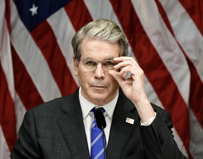 Treasury Secretary Bessent adjusts his glasses in front of an American flag, likely speaking about the CLARITY Act's progress in the Senate.