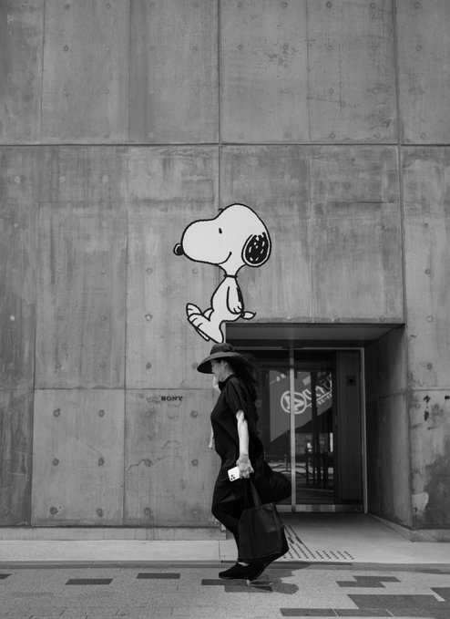 A black-and-white photo shows a woman in a hat and dress walking past a concrete wall featuring a mural of Snoopy sitting above a doorway.