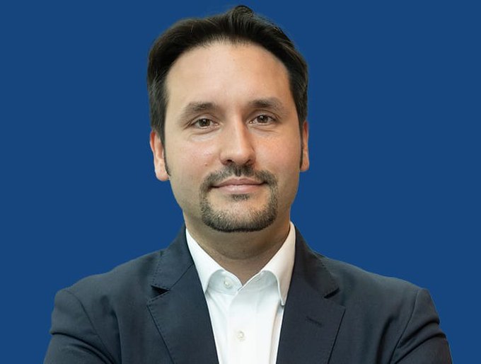 The image features a man wearing a suit jacket sitting against a solid blue background. He has his hands resting behind his head and looks directly at the camera with an expression of concern or contemplation. There is also text in the image, which reads "bitwise's André Dragosch sees up to 20% more downside for Bitcoin, with a worst case scenario placing the bottom near $48,000." This suggests that there might be some negative implications related to Bitcoin and its users' investments.