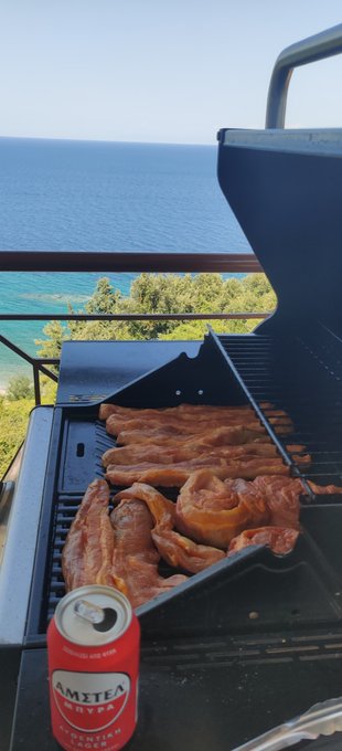 A photo showing strips of bacon cooking on a grill overlooking a beautiful ocean view, with a can of Amstel beer visible in the foreground.