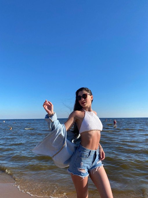 A young woman wearing a white halter bikini top, denim cutoff shorts, and holding a denim jacket, posing on a beach with clear blue sky and ocean in the background.
