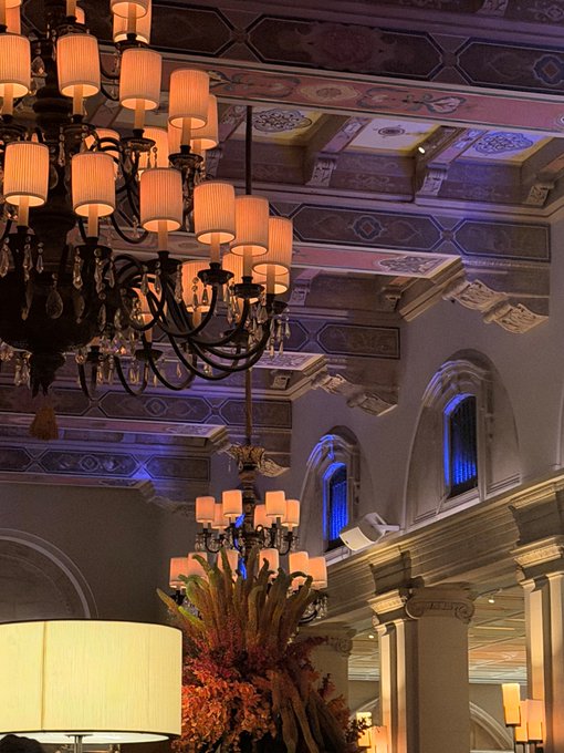 An interior view of an ornate, luxurious lobby or hall featuring elegant multi-tiered chandeliers with cream lampshades, decorative coffered ceilings, and classical architectural details.