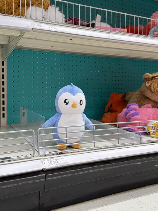 A blue and white penguin plush ("Pengu") priced at $10.99 sits alone on a Target store shelf, surrounded by other stuffed animals on a teal pegboard display. The image visually supports the caption's observation about the plush selling through quickly, as this location appears to have only one Pudgy Penguin left.