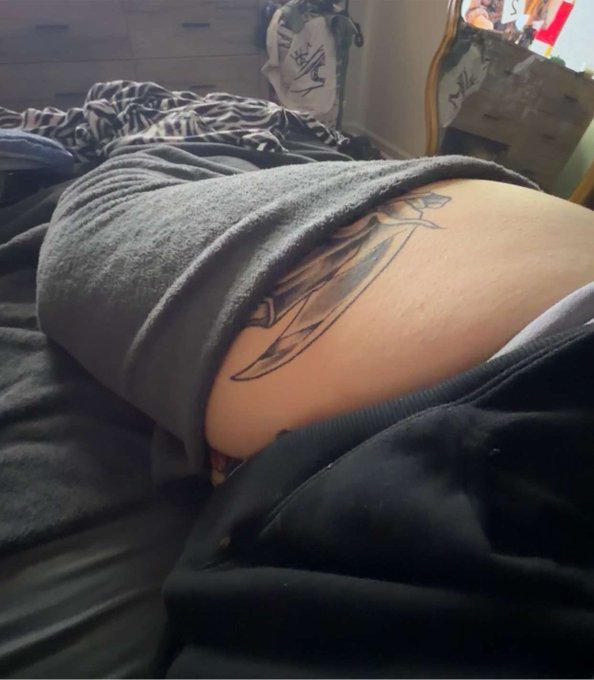 A person lying face-down on a bed, with a tattoo partially visible on their lower back/hip area featuring a banner and script lettering.