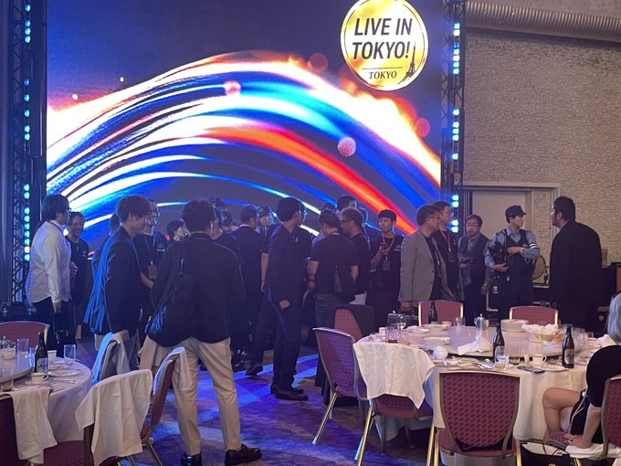 Attendees gather at a "LIVE IN TOKYO!" Bitradex Tokyo event, with uniformed police officers present among the crowd near round banquet tables set with wine bottles and glasses. The scene reflects the post's mention of police presence causing a stir at the cryptocurrency event.