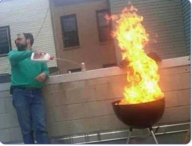 A bearded man in a green sweater pours lighter fluid onto an already flaming charcoal grill, sending a towering column of fire into the air. The image humorously illustrates the post's "Keep cooking" reference, capturing Charles Hoskinson's grill session in dramatic, over-the-top fashion.