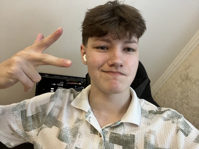 A selfie of a young boy making a peace sign, wearing a white patterned shirt and an earbud, with a computer monitor visible in the background.