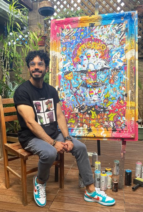 A smiling young man with curly hair and glasses, wearing a black graphic t-shirt and teal sneakers, sits on a wooden chair in front of a large, colorful graffiti-style painting on an easel, with spray paint cans arranged at his feet.