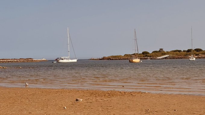 A coastal scene shows three sailboats anchored on calm water near a rocky shoreline, with a sandy beach in the foreground.