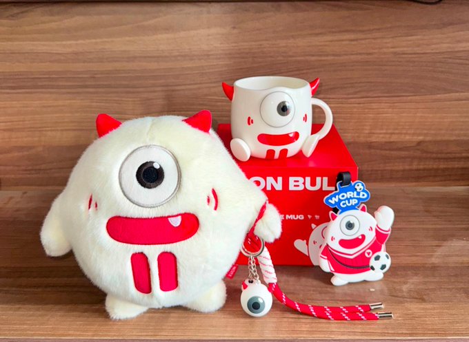 A merchandise display featuring a plush toy, mug, and lanyard with a one-eyed red monster character, alongside 'WORLD CUP' branding and a 'LION BULL' box.