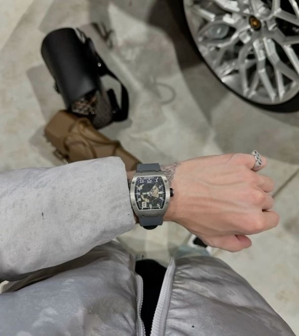 A first-person photo showing a wrist wearing a luxury skeleton watch with a blue strap and a silver ring, with a designer handbag and a Lamborghini wheel visible in the background.