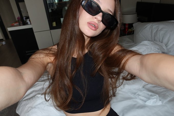 A selfie of a woman with long brown hair wearing black sunglasses and a black top in what appears to be a bedroom setting.